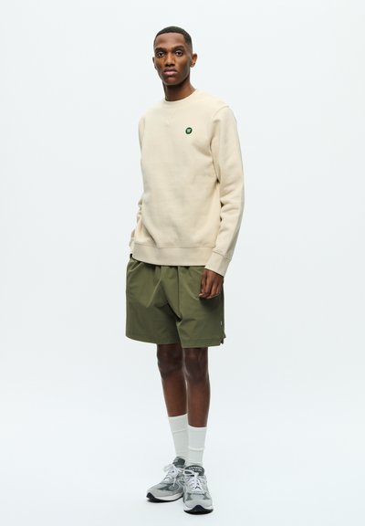 Beige Sweatshirt mit kleinem grünem Logo, kombiniert mit lockeren olivgrünen Shorts, weißen Crew Socken und grauen Trainingsschuhen vor einem schlichten Hintergrund.