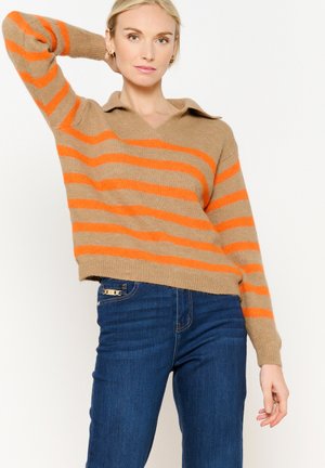 Pull beige avec des rayures orange, en col en V et avec un col. Porté avec un jean bleu. Texture douce, coupe décontractée et manches longues.