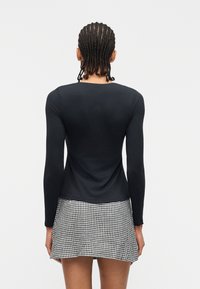 Άτομο με πλεξουδάκια, απεικονιζόμενο από πίσω, φοράει μαύρο μακρυμάνικο κορμάκι και μαύρη-λευκή καρό φούστα, πάνω σε απλό φόντο.