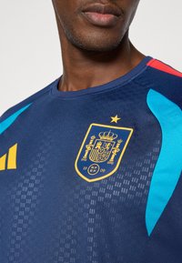 Vīrietis valkā tumši zilu sporta krekli ar dzeltenu Spānijas nacionālās komandas emblēmu un zilām un sarkanām plecu detaļām.