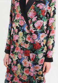 Blazer fleuri avec un fond noir, orné de grandes roses roses et rouges, rehaussé de fleurs bleues. Deux boutons noirs et revers noirs contrastants.