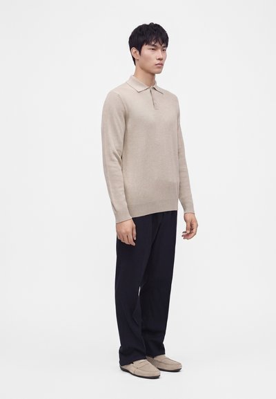 Homme debout portant un polo beige à manches longues, un pantalon noir et des chaussures beige type slip-on sur un fond blanc uni.