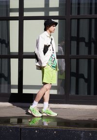 Leichte weiße Jacke mit schwarzen Akzenten, grünes Grafikshirt, neongrüne Shorts, weiße Socken und leuchtend grüne Sneakers. Schwarzer Bucket Hat.