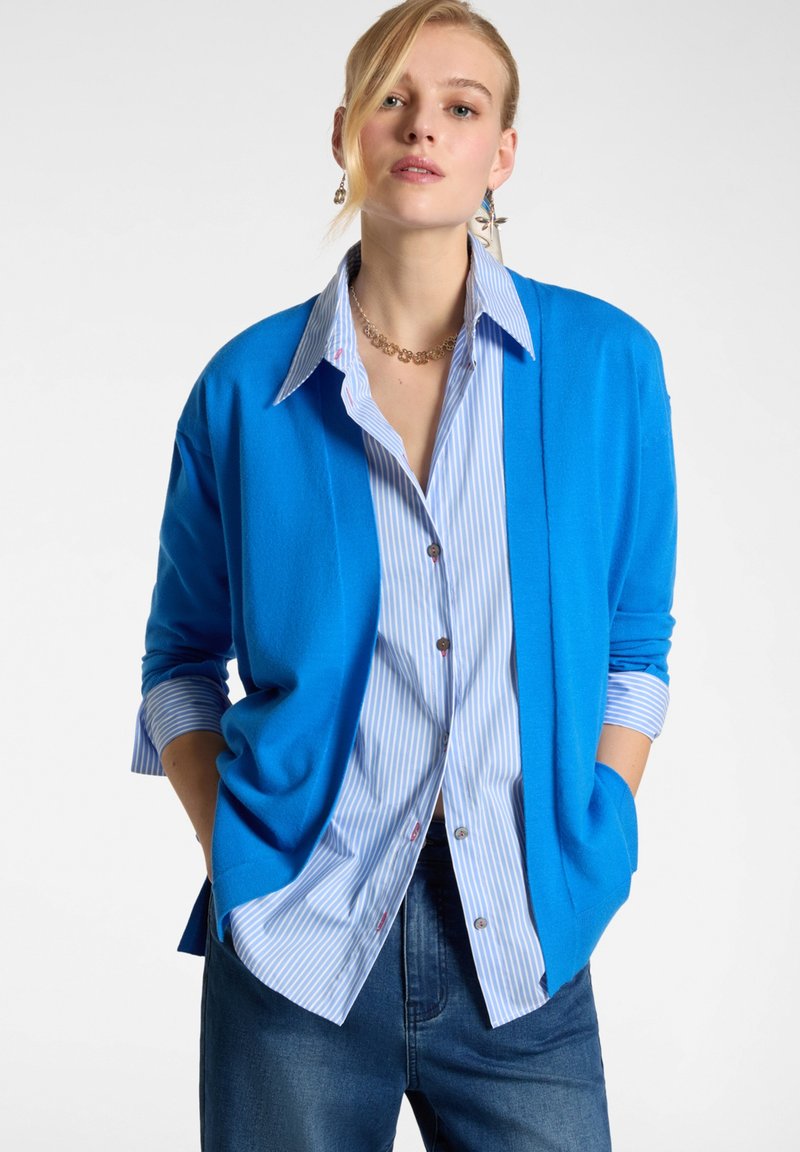 Cardigan blu aperto realizzato in un materiale morbido e leggero. Presenta maniche lunghe e una vestibilità comoda, indossato sopra una camicia button-up a righe blu chiaro.