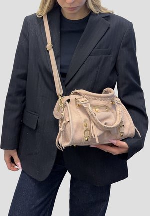 Donna con blazer nero a righe sottili che tiene una borsa a tracolla beige in suede con borchie dorate e tracolla sulla spalla.