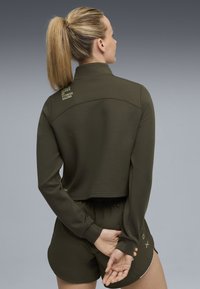 Giacca corta verde oliva con colletto alto e dettaglio del logo sulla schiena, abbinata a pantaloni corti coordinati con cuciture a contrasto e tasche.