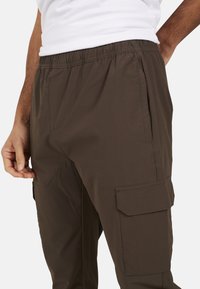 Ciemnobrązowe spodnie cargo z elastycznym pasem, kieszeniami bocznymi oraz ozdobnymi kieszeniami na udach. Gładka tekstura i luźny krój.