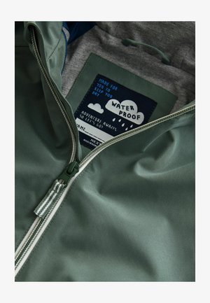 Veste imperméable verte avec une texture lisse, fermeture éclair, et un intérieur gris avec une étiquette imprimée et des graphismes ludiques.