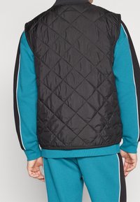 Svart vadderad väst med diamantmönster, med hög krage och ärmlös design, i kombination med en teal långärmad topp under.