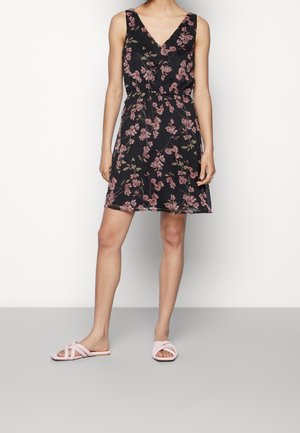 Frau trägt ein schwarzes ärmelloses Kleid mit rosa Blumen und hellrosa flachen Sandalen, steht vor einem schlichten weißen Hintergrund.