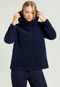 Kobieta poprawiająca wysoki kołnierz granatowej, teksturowanej wełnianej kurtki z przednimi kieszeniami naszytymi, nosząca pasujące ciemne spodnie.