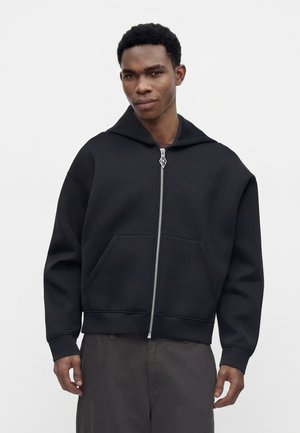 Jeune homme portant un sweat à capuche noir zippé avec des poches avant et un pantalon foncé, debout devant un fond clair uni.