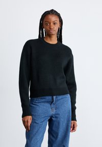 Črna pletena jopa z okruglim izrezom in rebrastim robom. Mehka tekstura in dolgi rokavi. Ujemita z svetlo modrimi jeans hlačami.