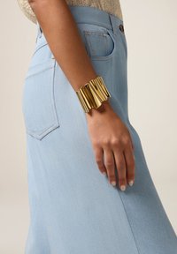 Frau trägt einen hellblauen Denim-Rock und ein großes, glänzendes goldfarbenes Armreif am Handgelenk, mit einem sichtbaren, beigefarbenen, strukturierten Oberteil.