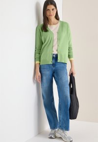 Femme aux longs cheveux foncés portant un cardigan vert clair, une chemise beige, un jean bleu et des baskets argentées, tenant un sac fourre-tout noir.