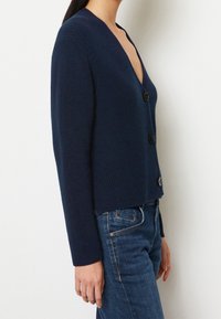 Marineblauer, gestrickter Cardigan mit tiefem V-Ausschnitt, langen Ärmeln und drei schwarzen Knöpfen vorne, kombiniert mit blauen Jeans.