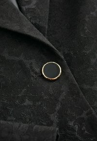 Black textured fabric with a floral pattern, featuring a round gold and black button. The design includes peaked lapels and a smooth finish.