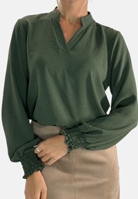 Long-sleeve, dark green blouse with a V-neck design, featuring textured fabric and gathered cuffs. Paired with a beige skirt.