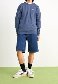 Niebieska bluza z okrągłym dekoltem i logo, w połączeniu z dżinsowymi szortami Bermuda. Obuwie to białe trampki z czarnymi akcentami oraz czarne skarpetki.