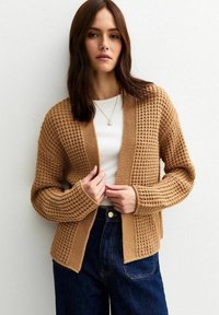 Cardigan a maglia in beige caldo, caratterizzato da un design testurizzato con apertura frontale e maniche lunghe. Abbinato a una maglietta bianca e pantaloni di jeans.