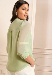 Blusa verde chiaro con maniche a sbuffo trasparenti, schiena arricciata e decorativi chiodini dorati lungo le maniche. Tessuto morbido, vestibilità comoda.