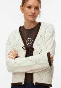 Cardigan en maille crème avec motifs en losange et accents en boules, présentant une bordure contrastante marron et une fermeture par lien au niveau de l'encolure.