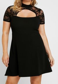 Robe noire avec des manches courtes en dentelle, un col haut, et une découpe à l'avant. Silhouette en A avec une texture de tissu lisse.