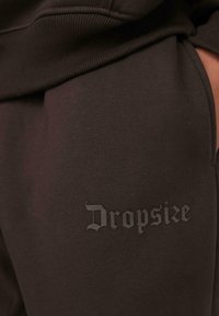 Close-up of dark brown sweatpants with ribbed waistband and "Dropsize" logo in gothic font on the thigh near a hand in the pocket.