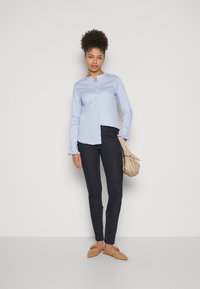 Camicia azzurro chiaro con maniche lunghe e polsini arricciati, abbinata a pantaloni scuri aderenti e scarpe basse beige, mentre tiene una borsa morbida beige.