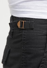 Svart tygbälte med en rektangulär bronsspenne, fäst vid byxor med en texturerad yta och horisontell ribbad mönstring, som visar fickdetaljer.
