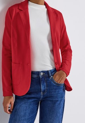 Henkilö pukeutuneena kirkkaan punaiseen bleiseriin valkoisen ribbineulotun topin ja sinisten farkkujen päällä, yksi käsi taskussa ja neutraali tausta.