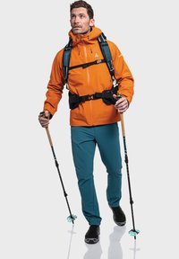 Orangene wasserdichte Jacke mit hohem Kragen, verstellbaren Trägern und türkisfarbenen Hosen. Ausgestattet mit Trekkingstöcken und schwarzer Fußbekleidung.