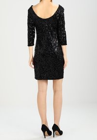 Svart paljettminikjol med trekvartsärmar, rundad rygg, åtsittande silhuett, texturerad yta, bärs med svarta högklackade skor.