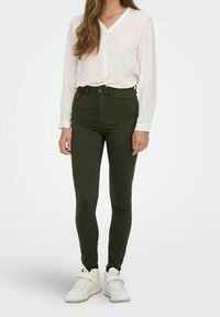 Jean skinny khaki taille haute, associé à une blouse blanche à boutons et des baskets blanches. Tissu lisse avec un détail minimal.