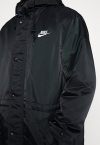 Veste Nike noire avec une finition lisse et brillante, boutons-pression sur le devant et une capuche avec cordon de serrage, affichant le logo Nike brodé sur la poitrine.