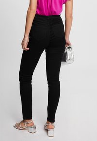 Jeans negros de cintura alta con una textura suave, que cuentan con bolsillos traseros y un diseño ajustado. Combinados con sandalias de tacón plateadas y un bolso metálico.