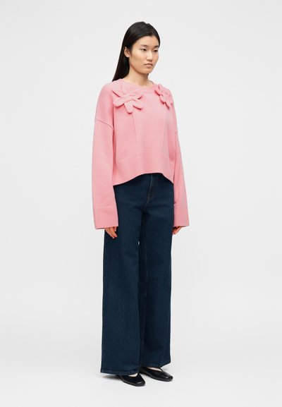 Ροζ cropped πουλόβερ με εφαρμογές λουλουδιών, μανσέτες με ρίγα και άνετη γραμμή, συνδυασμένο με παντελόνι σε σκούρο τζιν με φαρδιά πόδια και μαύρα παπούτσια.