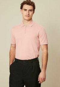 Polo shirt roz deschis cu fermoar la gât și mâneci scurte, asociat cu pantaloni negri cu un model texturat. Design simplu cu o croială lejeră.