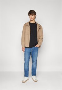 Tan jas met knoopsluiting, zwart T-shirt, blauwe jeans en witte sneakers met beige accenten. Staande tegen een eenvoudige achtergrond.