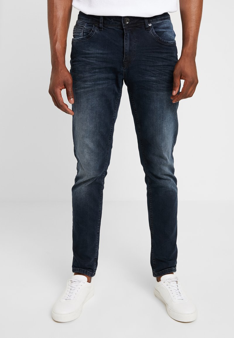 Donkerblauwe denim jeans met een slim fit, voorzien van een vervaagde textuur en subtiele pocketdetails, gecombineerd met witte sneakers.