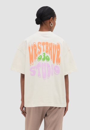 Kobieta z ciemnymi włosami związanymi w kok, ubrana w luźną białą koszulkę z kolorowym falistym napisem „WHSTBHVR 030 STUDIO” na plecach, beżowe spodnie.