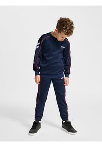 Mörkblå träningsoverall i mjukt tyg med röda sidostråk. Inkluderar en tröja med rundad hals och byxor med mudd. Svarta sportskor.