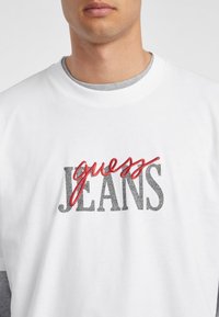 Sudadera blanca con la inscripción "guess JEANS" en letras texturizadas negras y rojas. Diseño de cuello redondo, fabricada en un tejido suave, con el interior del collar en gris visible.