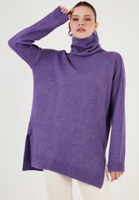 Vijolična pulover-turtleneck iz mehkega materiala, s sproščenim krojem, rebrastimi manšetami in obliko spodnjega dela, ki je višje spredaj in nižje zadaj.