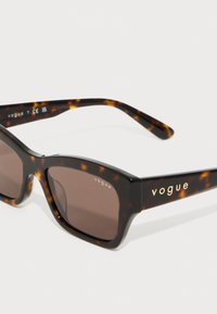 Ochelari de soare maro cu modele tortoise, cu lentile rectangulare. Ramele au un finisaj lucios, iar pe lateral este gravat numele brandului "vogue".