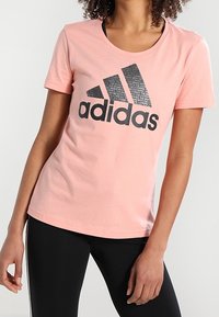 Różowy bawełniany t-shirt z dużym czarnym logo Adidas na przodzie. Krótkie rękawy i okrągły dekolt, gładka faktura.
