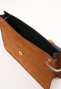 Hnědá semišová taška přes rameno s černým lemem, která má magnetické zapínání a zlaté kovové doplňky. Uvnitř se nachází zipová kapsa.