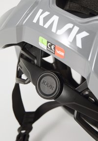 Casque de cyclisme KASK en gris mat avec visière noire ajustable et molette. Comprend des étiquettes de sécurité et des sangles de menton sécurisées pour un bon ajustement.