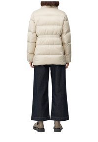 Osoba oblečená v béžovej pérovke, tmavých širokých džínsach a béžových robustných topánkach, stojí chrbtom na bielom pozadí.