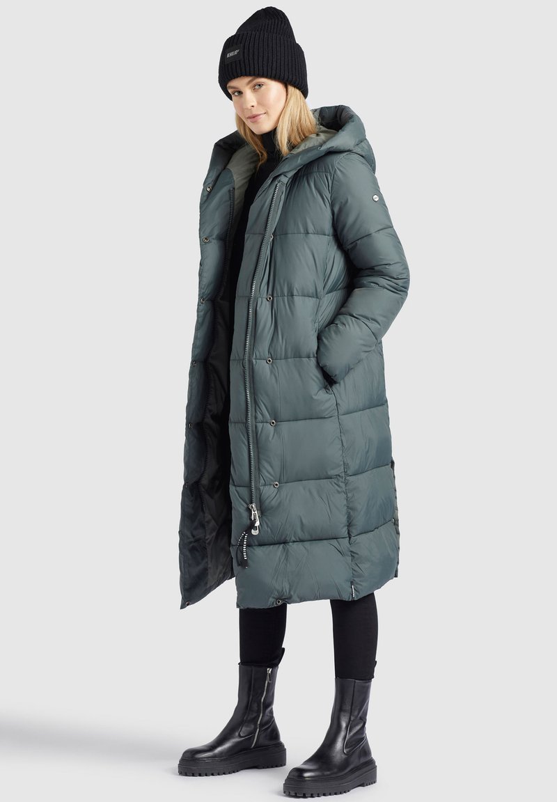 Frau trägt einen langen, tealfarbenen, gepolsterten Mantel mit Kapuze, schwarze Mütze, schwarze Hose und schwarze, klobige Stiefel. Sie steht vor einem schlichten grauen Hintergrund.